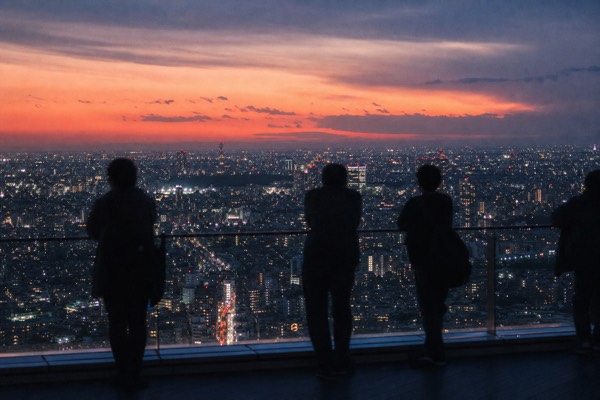 Tokyo skyline at sunset from a high open rooftop, sky in bands of coral and indigo over the city lights