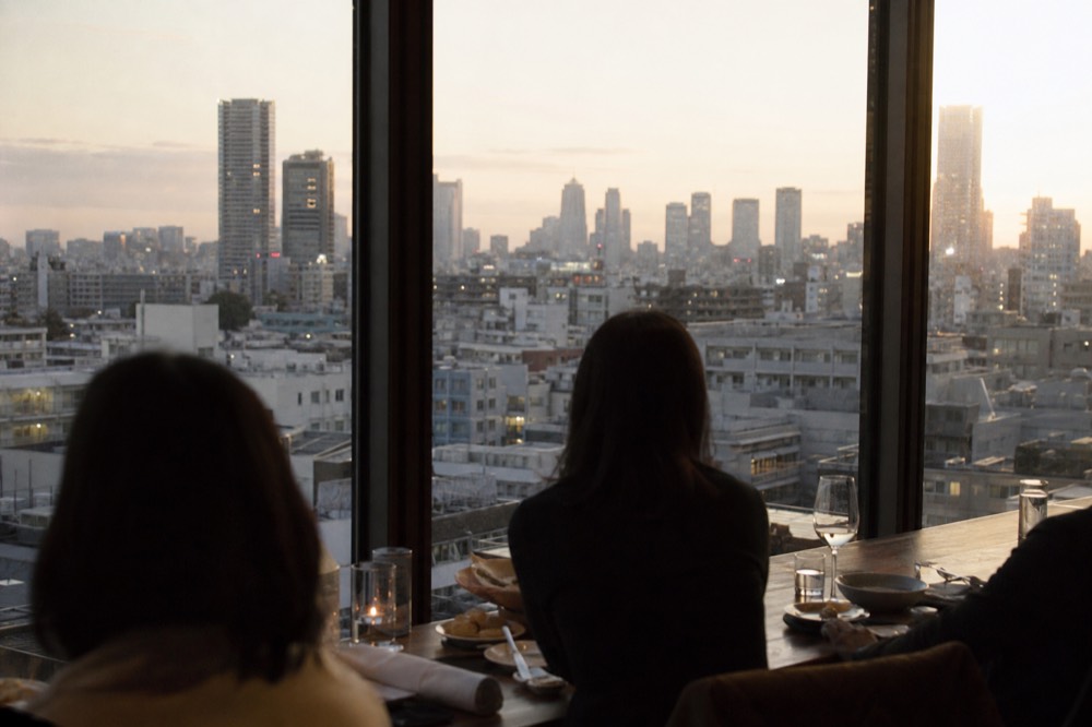Diners at a window table on a mid level restaurant floor looking out over Tokyo rooftops in late afternoon light