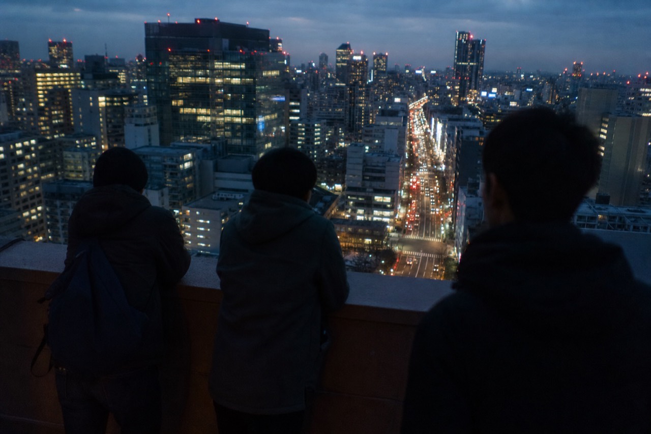 Open rooftop parapet high above a Tokyo intersection at dusk with visitors looking down at traffic and lit crossings far below