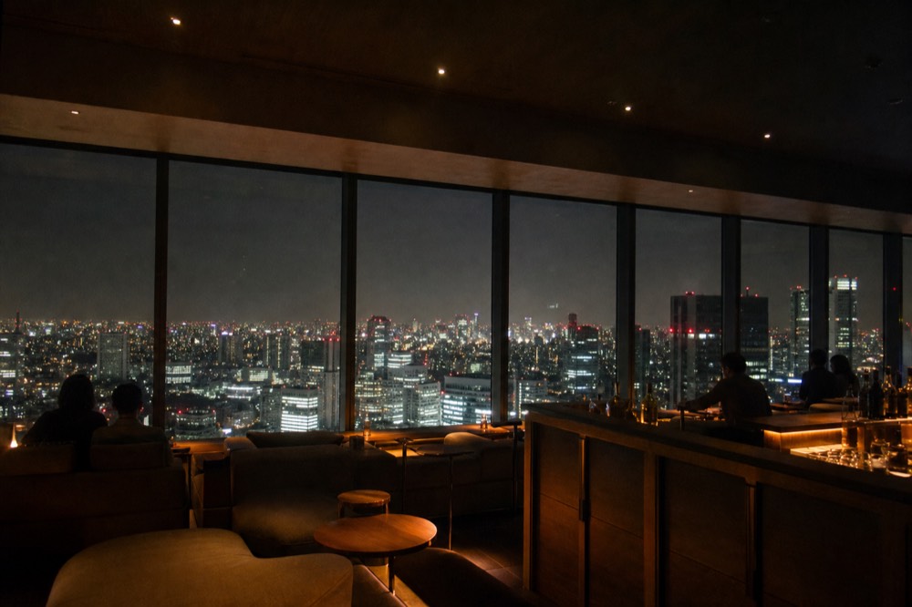 Warm indoor observation lounge on a high floor with low seating, a drinks counter and a wide night city view through tall glass