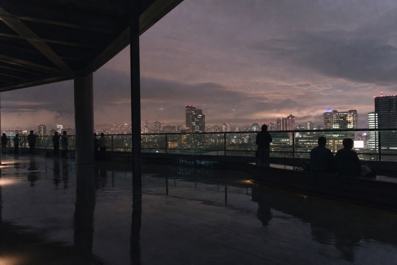 Visitors spread out along the low railing of an open rooftop deck at dusk, some seated, city lights coming on below