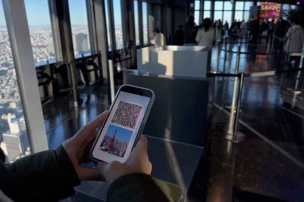 Close view of hands holding a phone with a timed entry ticket screen while standing in a busy Tokyo tower lobby