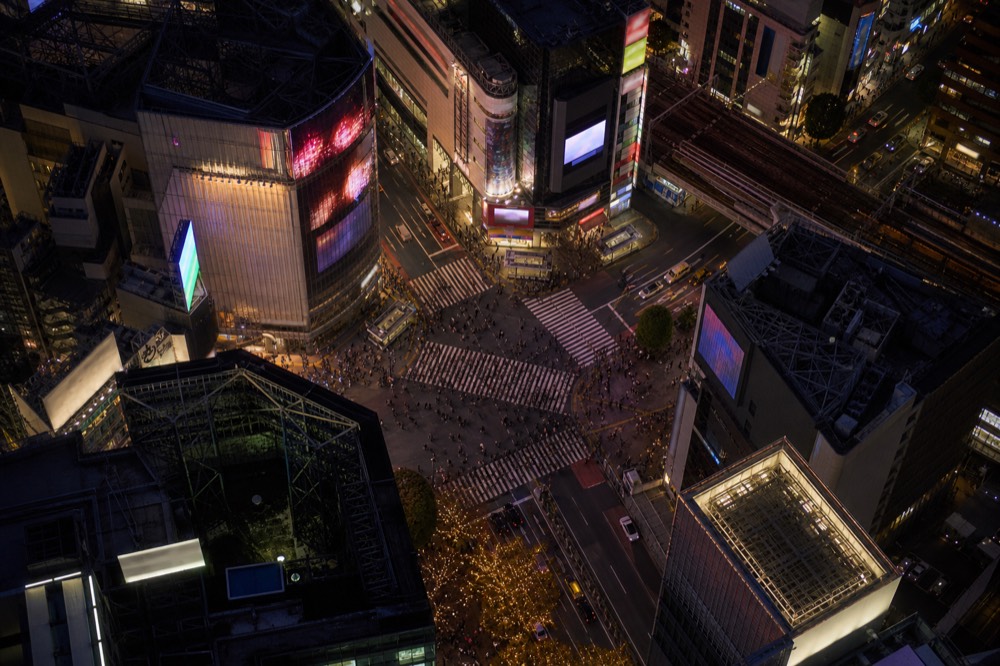 Aerial view straight down onto a busy pedestrian scramble crossing surrounded by illuminated advertising screens and rooftops