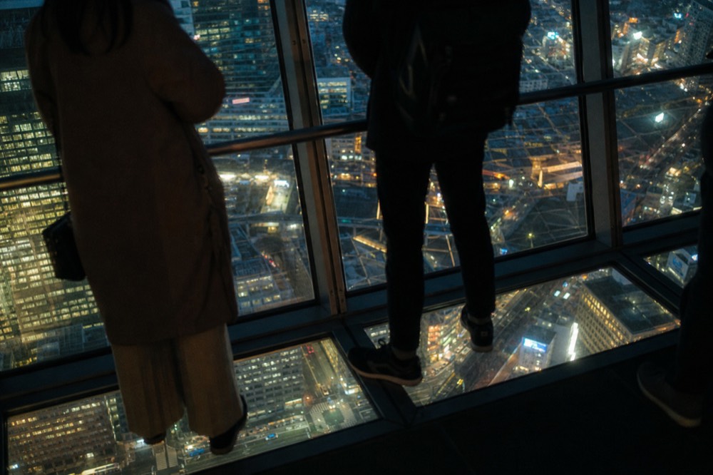 Visitors standing on a glass floor panel high inside a circular observation level looking down at streets far below