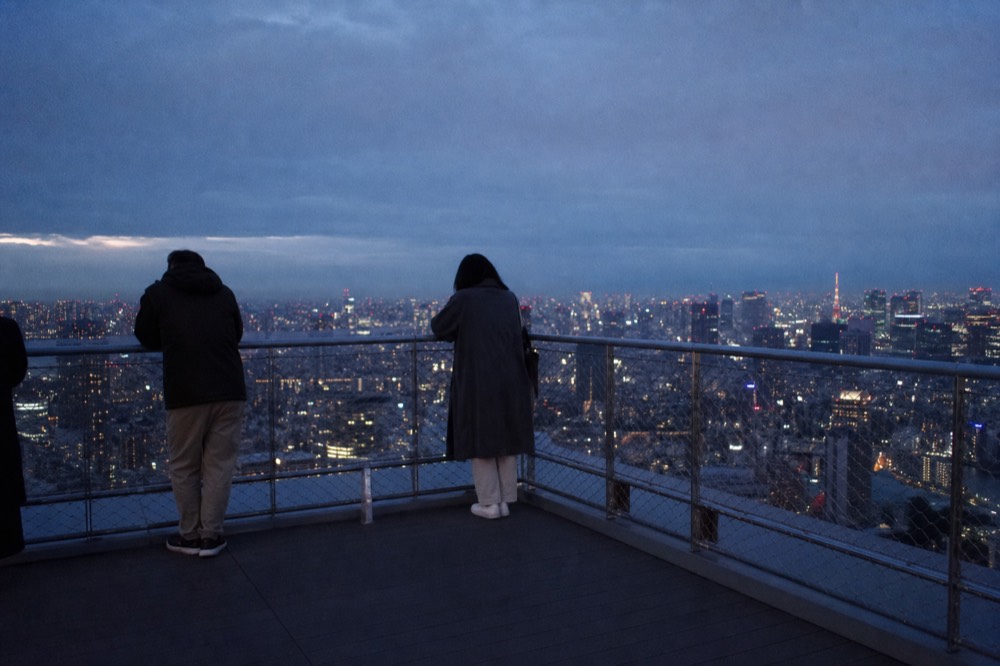 Open rooftop viewing area at the top of a Tokyo tower at dusk with visitors at the corner railing and city lights below