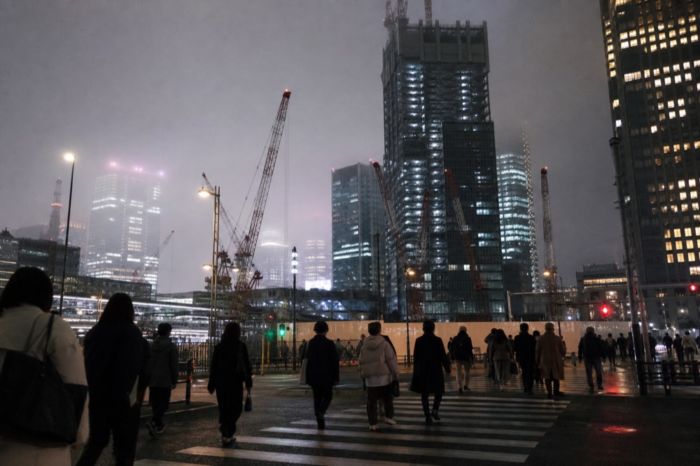 Construction cranes and half-finished towers around a Tokyo station plaza with pedestrians crossing in the foreground