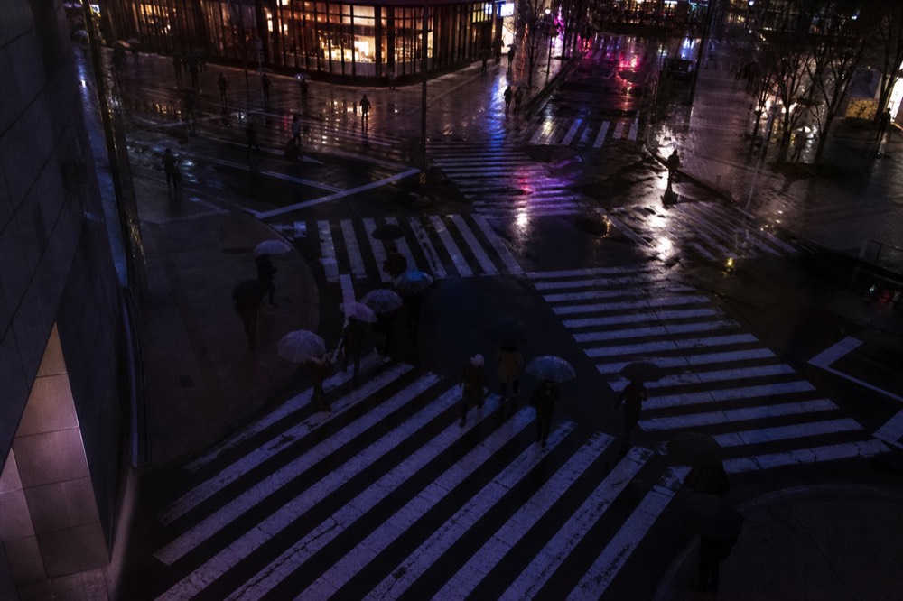Overhead view of pedestrians crossing diagonally across a scramble intersection with white markings on wet asphalt