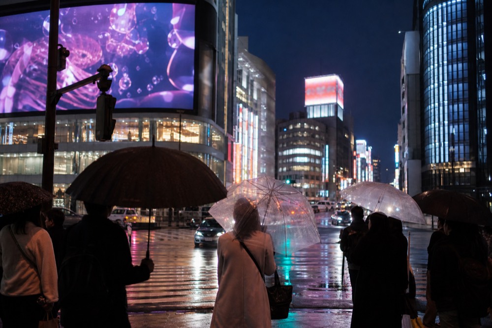 Large video screens and neon signage towering over a Tokyo intersection at dusk with crowds waiting at the kerb