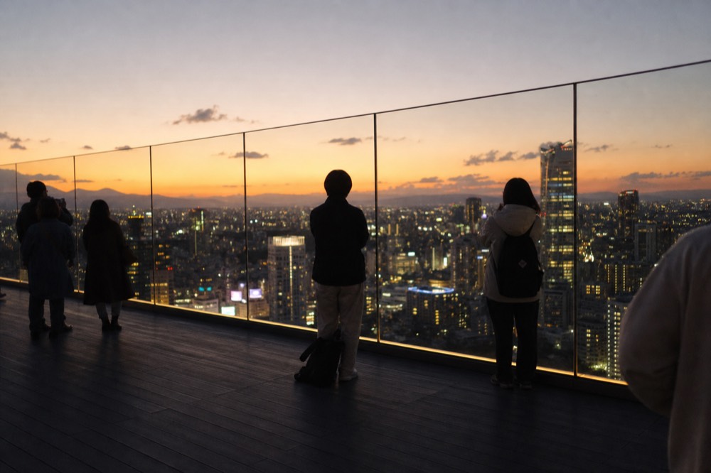 Open-air rooftop observation deck at dusk with visitors along a glass parapet facing a wide western horizon over a lit city