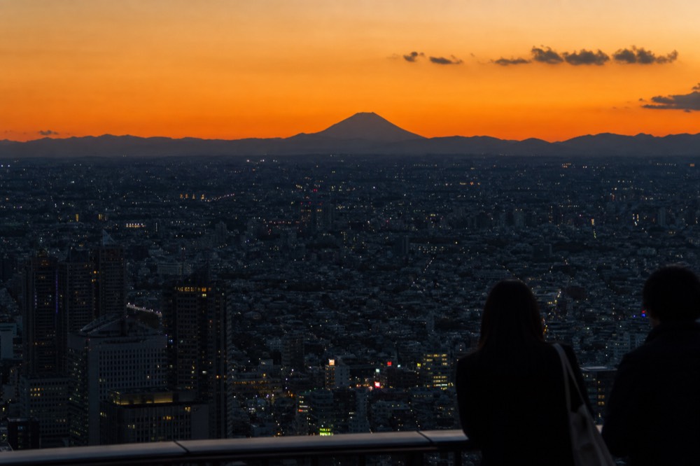 Backlit volcanic cone silhouetted against a deep orange sunset sky above a dark layered city horizon
