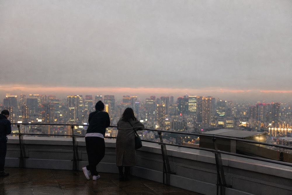 Visitors standing at the western railing of a high open rooftop scanning a distant horizon at golden hour