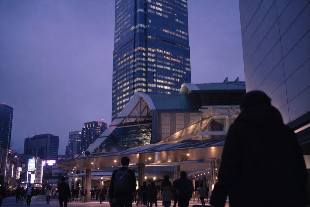 Tall glass tower rising directly above a busy railway station concourse in Tokyo, seen from street level at dusk