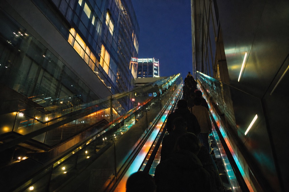 Long glass-sided escalator rising through a dark tunnel lit with strips of coloured light, silhouetted riders facing upward