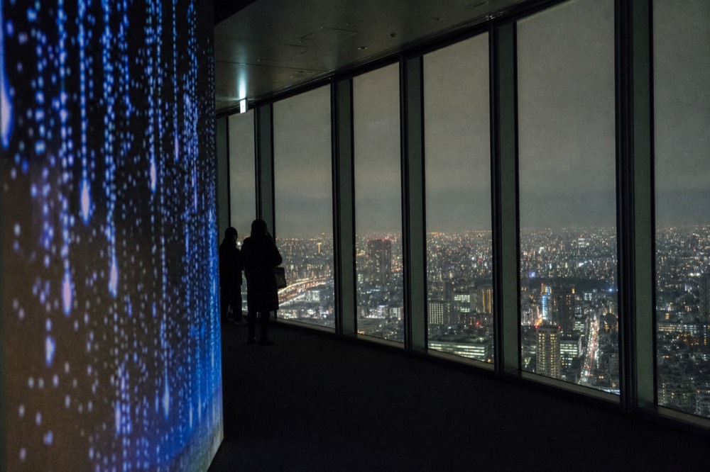 Dim indoor observation gallery with glowing digital art panels along one wall and a wide night cityscape through full-height windows
