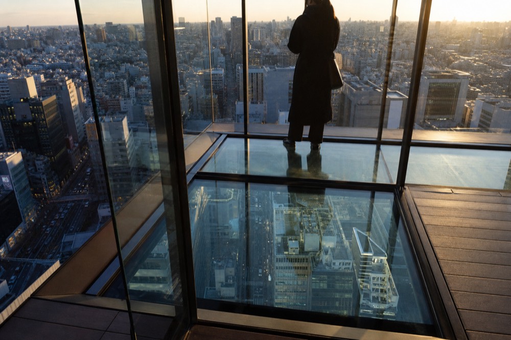 Glass-floored corner platform at the edge of a rooftop deck with the city street grid visible far below through the panel