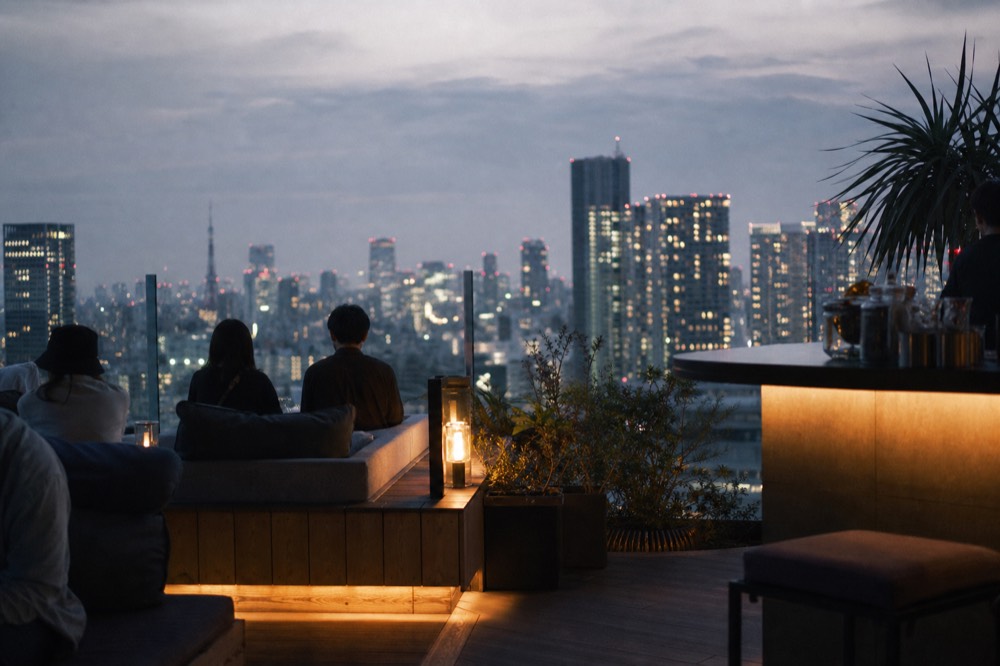 Low sofa seating and a drinks counter set up on an open rooftop at dusk with the lit skyline stretching behind