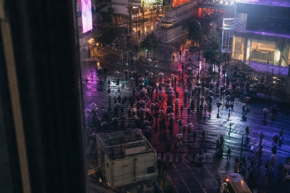 Pedestrian scramble crossing seen from a second floor window with crowds converging under neon screens at dusk
