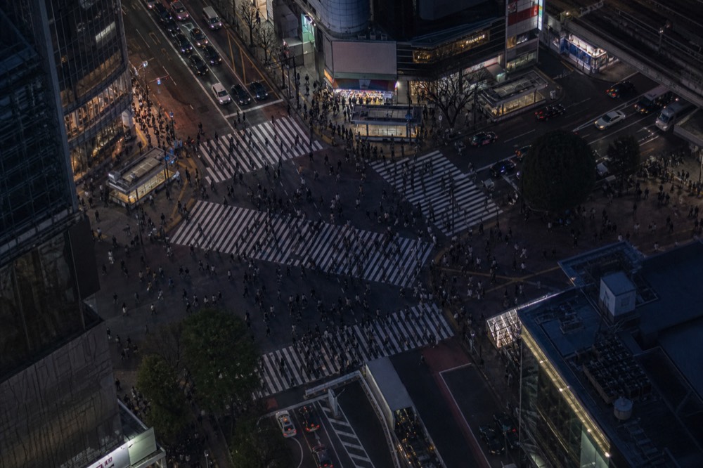 Aerial view straight down onto a scramble intersection at night with dense crowds crossing in every direction under white road markings
