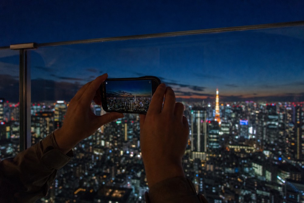 Close view of hands holding a phone pressed against a clear glass parapet with a lit city skyline beyond at night
