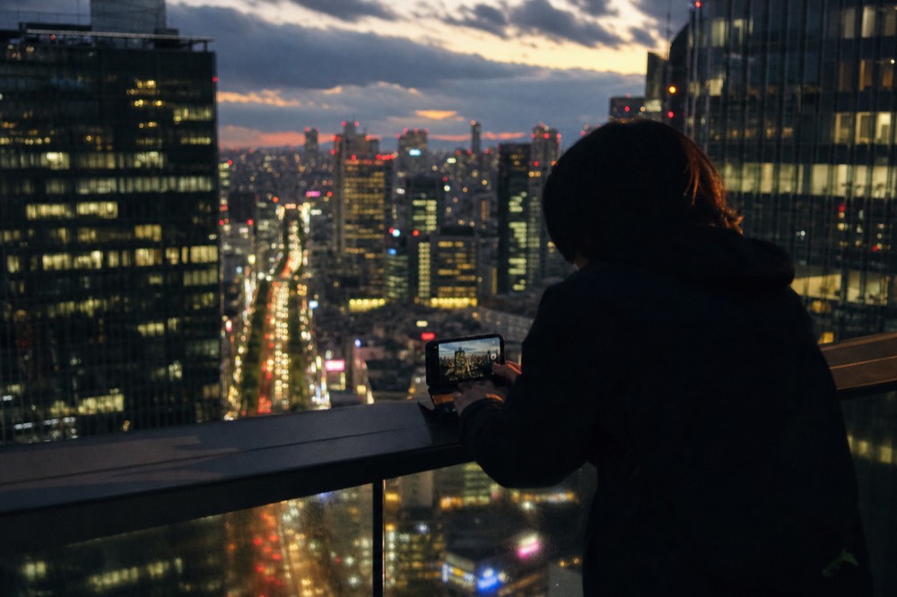 Visitor bracing a phone against a rooftop railing to photograph glowing streets and towers far below after dark