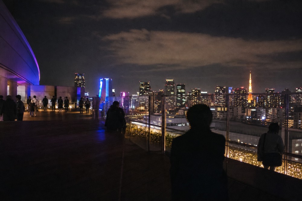 Coloured light sequence washing across an open rooftop deck at night with visitors silhouetted against a glowing city