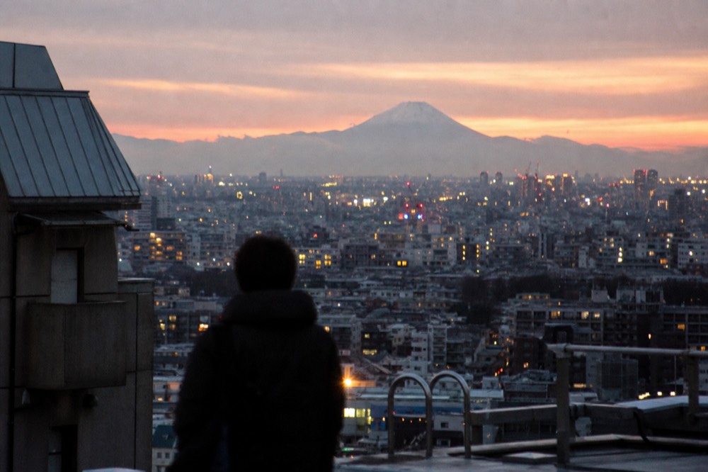 Low winter sun dropping behind a distant snow-capped volcano seen across a wide city skyline from a high open rooftop