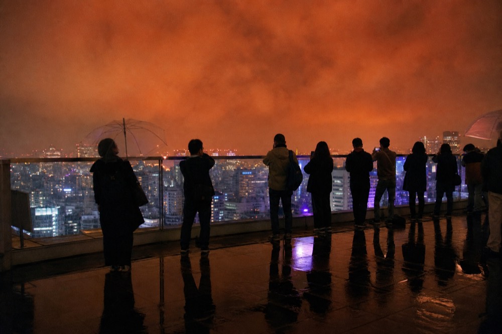 Dense line of visitors waiting at a glass corner railing on a rooftop deck while the sky behind them turns deep orange
