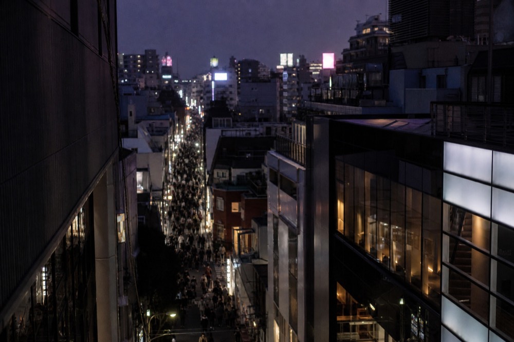 Crowded Tokyo shopping street at dusk seen from above, dense pedestrian flow between illuminated storefronts and vertical signage