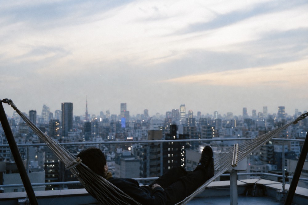 Visitor lying back in a suspended rope hammock on a high rooftop with only open sky and distant towers above
