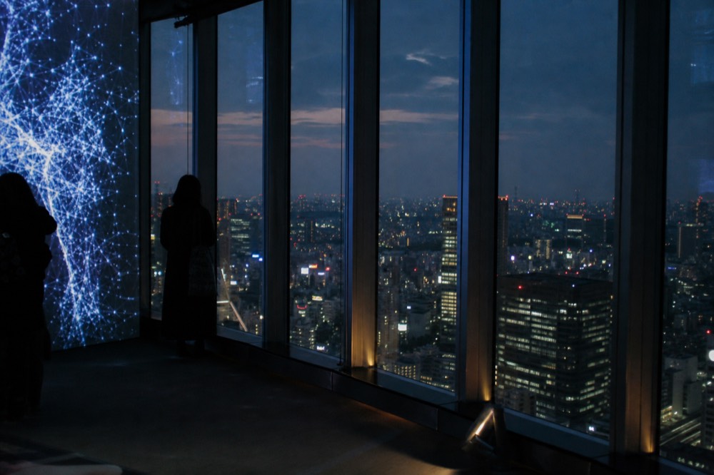 Indoor observation gallery with dark glass walls, glowing digital art panels and visitors watching the city lights beyond