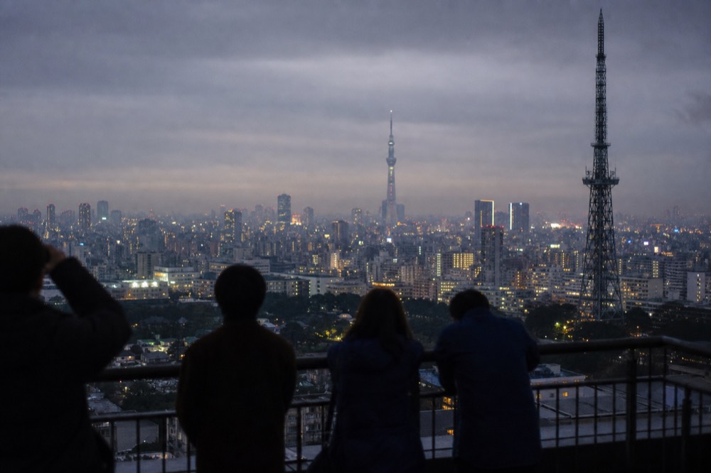Tokyo skyline at blue hour seen from a high vantage point with a tall lattice tower and a distant broadcasting tower on the horizon