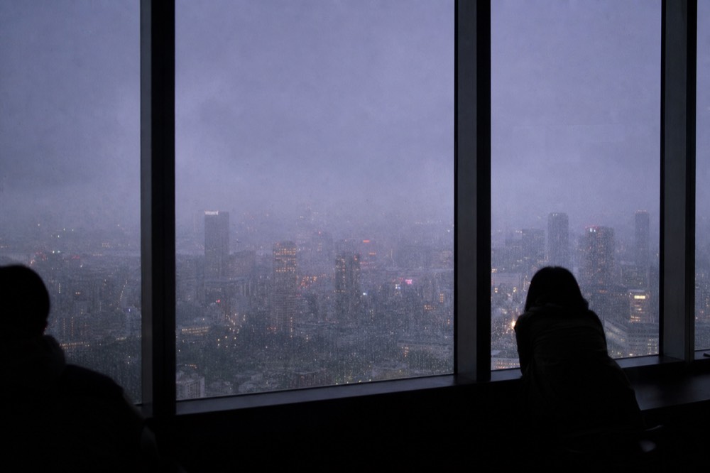 Rain streaked window on a high indoor observation floor with a grey city barely visible through low cloud outside