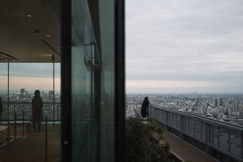 Split view of Tokyo from height, a glass-walled indoor observation level on one side and an open rooftop railing on the other, city stretching beyond both