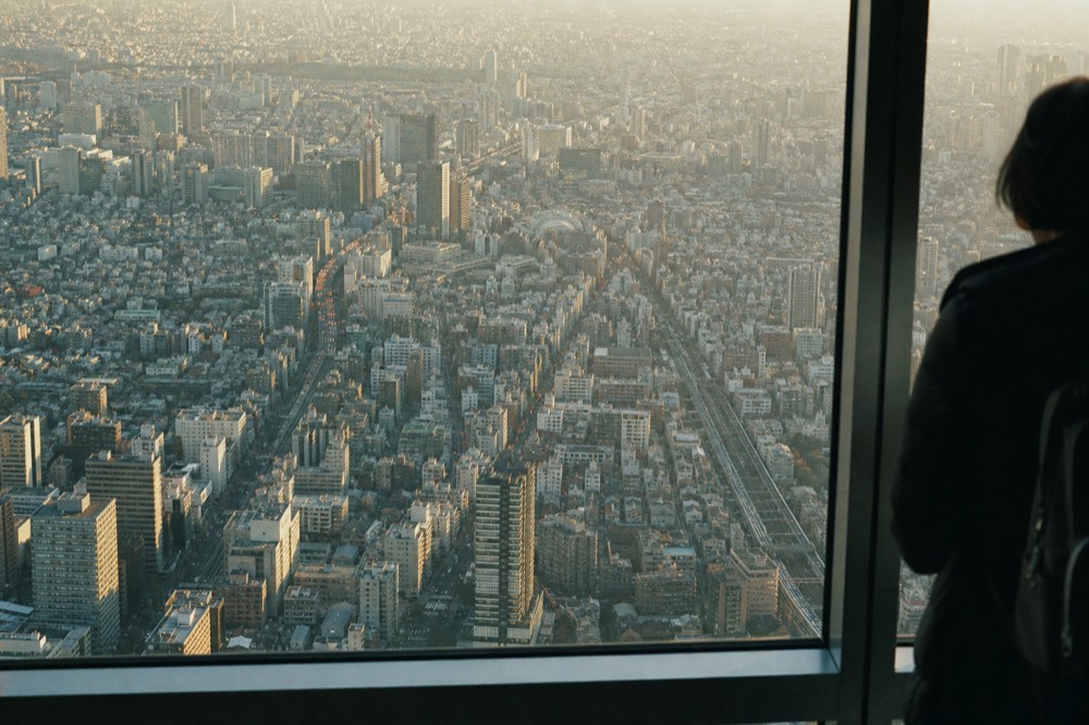Aerial view of dense Tokyo rooftops and rail lines seen from a great height, individual streets reduced to fine pattern under afternoon haze
