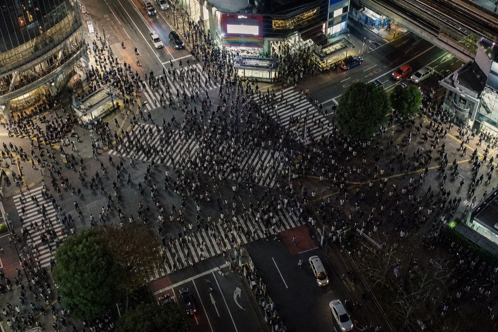 Looking down on a busy scramble intersection from directly above at night, crowds crossing in every direction under bright screens and street lights