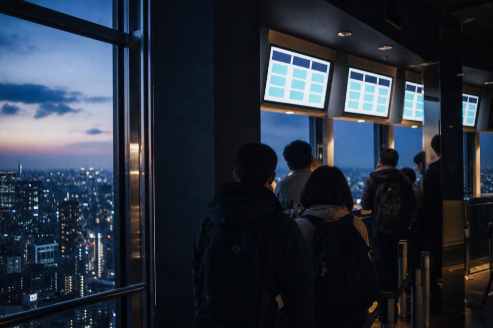 Ticket counter area on an upper floor of a Tokyo tower, digital time-slot displays above a short queue of visitors with backpacks