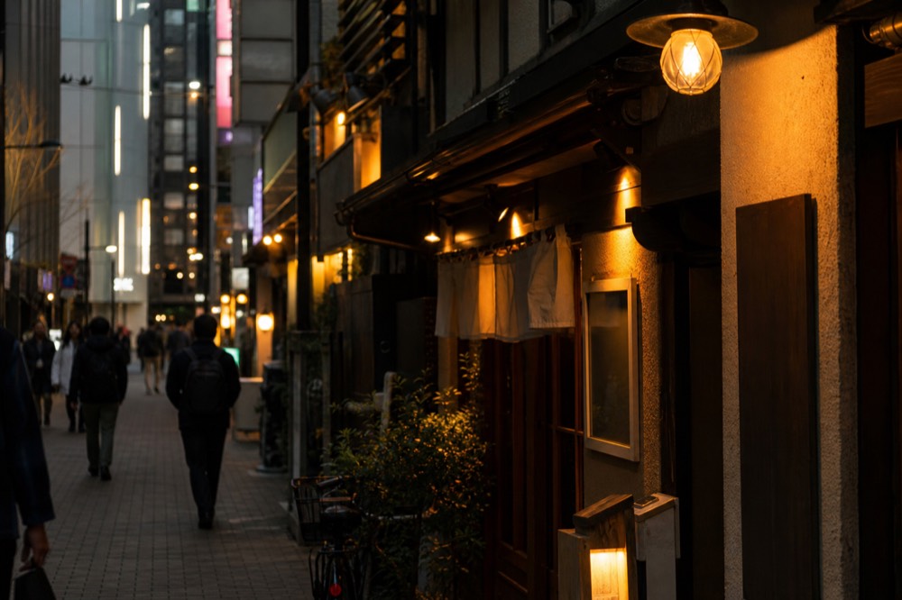 Narrow pedestrian shopping street lined with vertical illuminated signs and small restaurant entrances in the evening