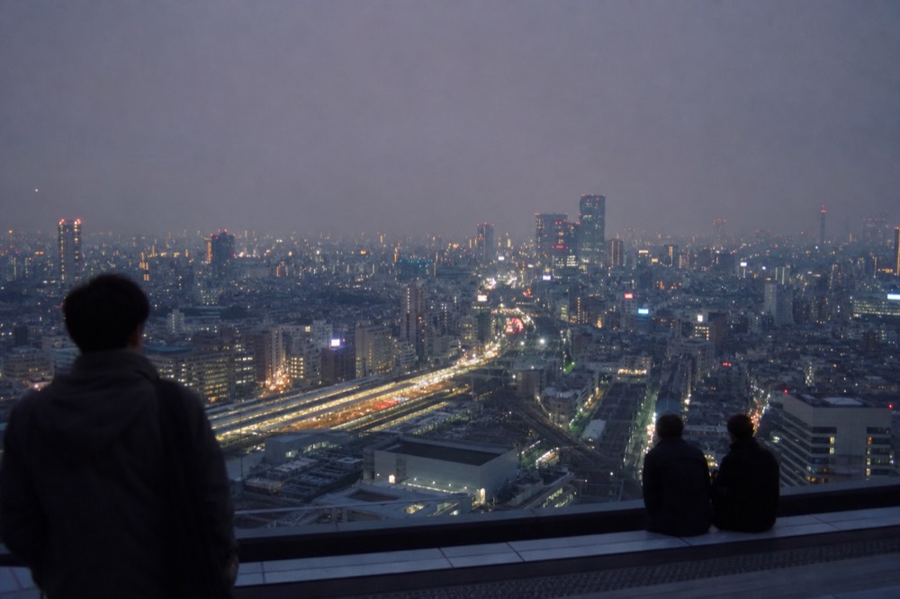 City district seen from a high open rooftop at blue hour with lit streets radiating out from a station complex