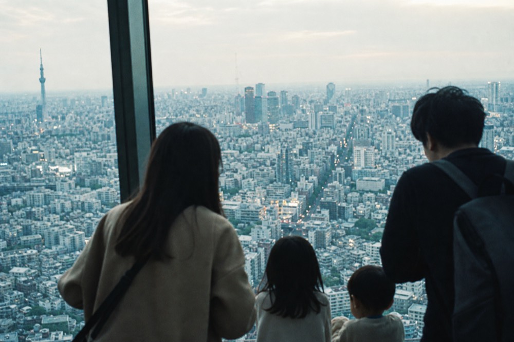 Family with two children at a tall observation window looking down on a Tokyo cityscape in late afternoon light
