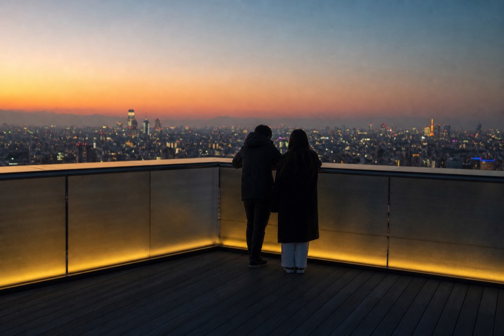 Open rooftop viewing deck at dusk with visitors at the corner railing and a city skyline turning from orange to blue behind them